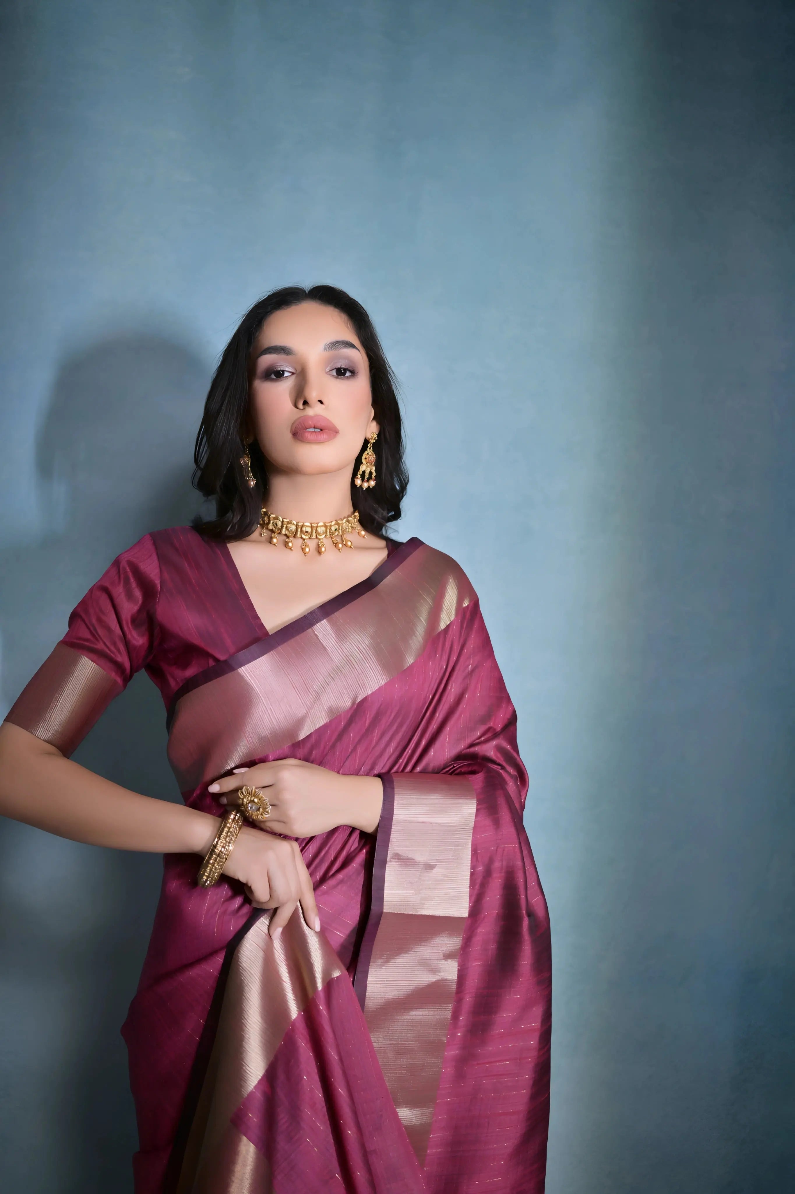 A woman models a striking Wine Red Banarasi Raw Silk Saree. The saree body features subtle woven stripes and is framed by an extremely wide, luxurious metallic gold Zari border. She wears a matching wine-colored blouse and traditional gold Kundan jewelry.